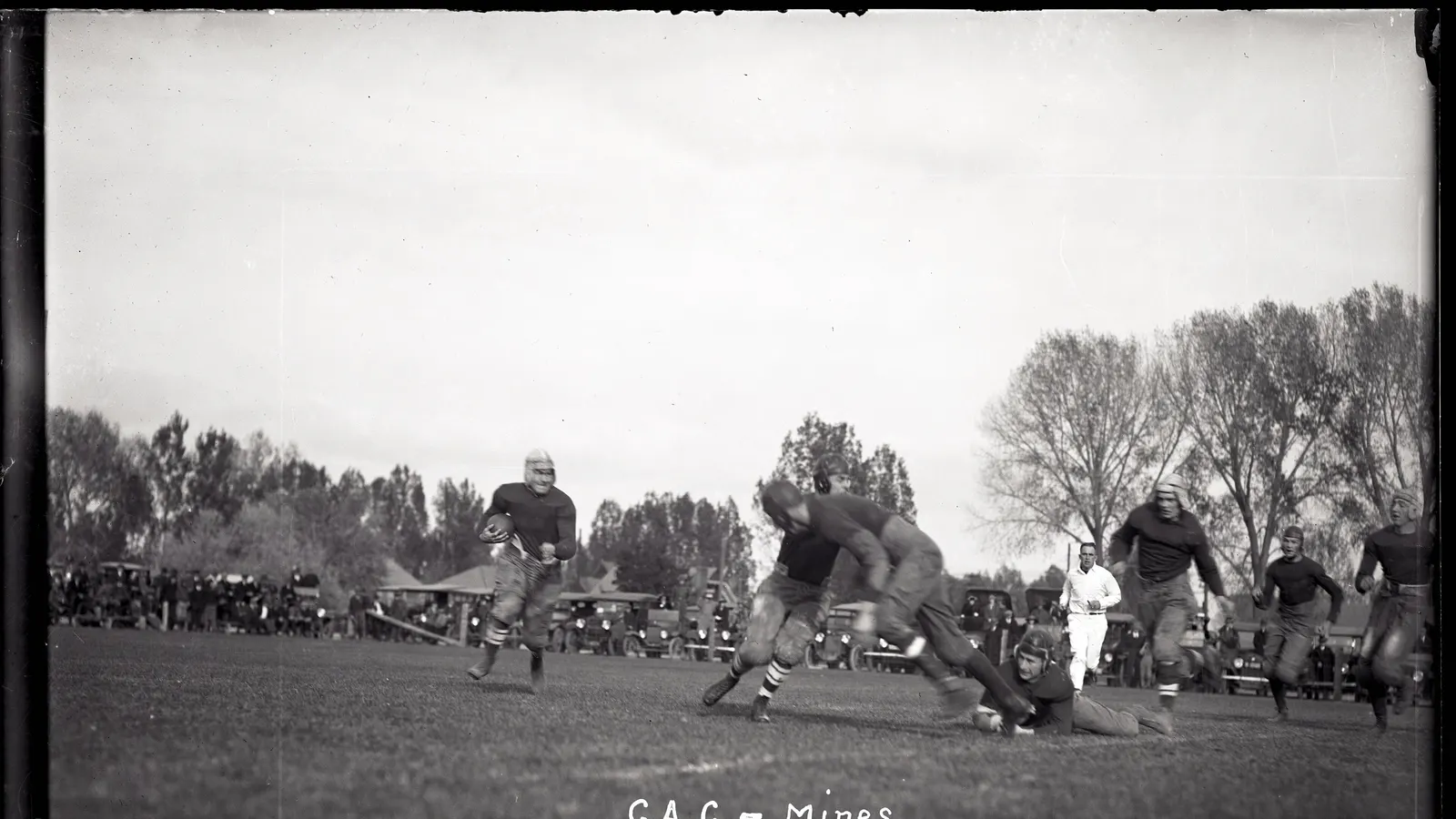 Joueurs d'un match universitaire des années 1920 en course avec le ballon, spectateurs dans des voitures garées